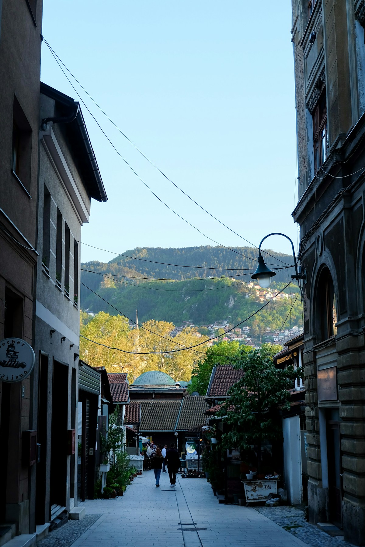 Narrow cobblestone street in the old town of Sarajevo with a mosque minaret and green hills behind